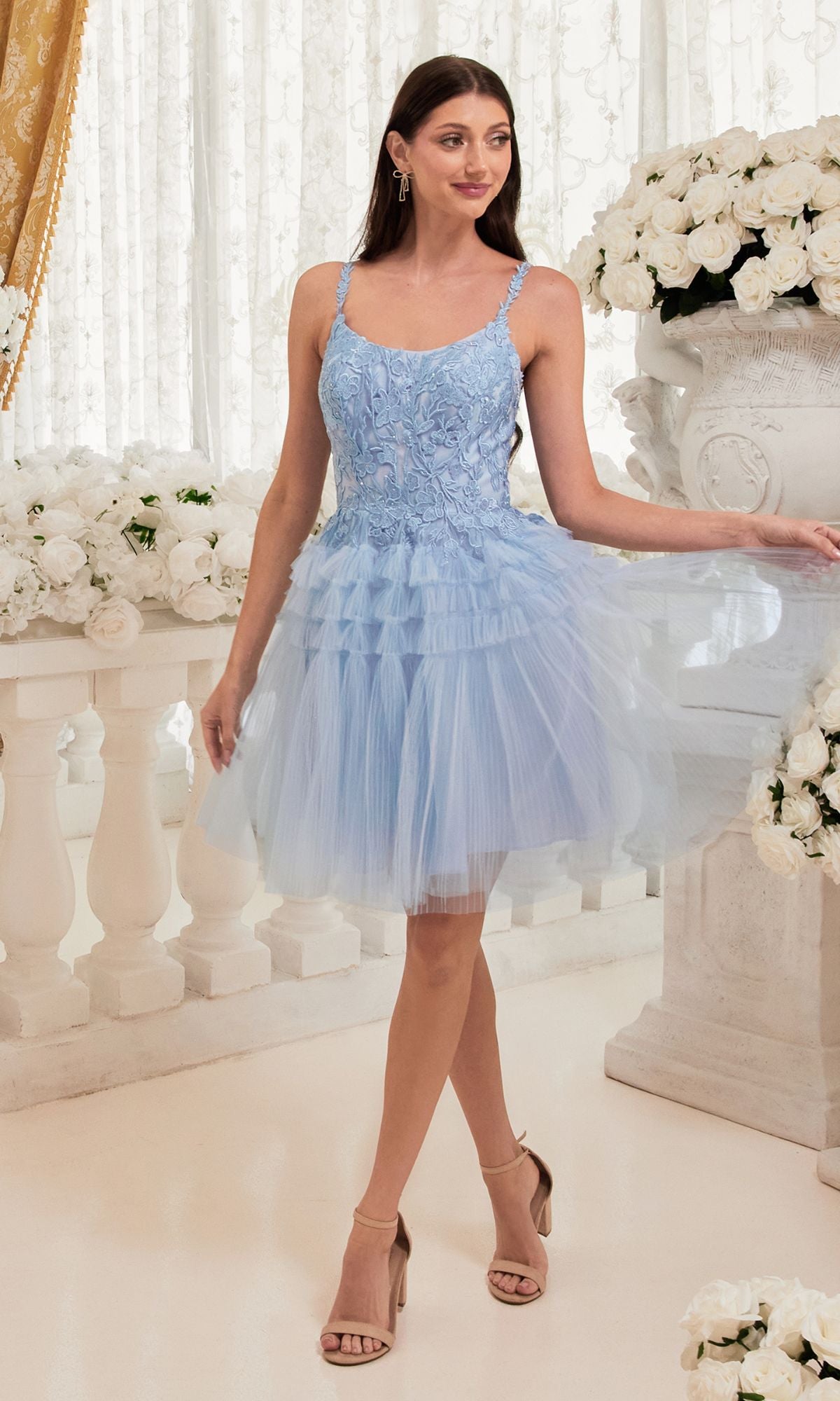 A woman in the Lace-Bodice Tiered-Skirt Short Prom Dress 9310, featuring a light blue lace bodice and thin straps, stands indoors surrounded by white floral arrangements and elegant classical decor.